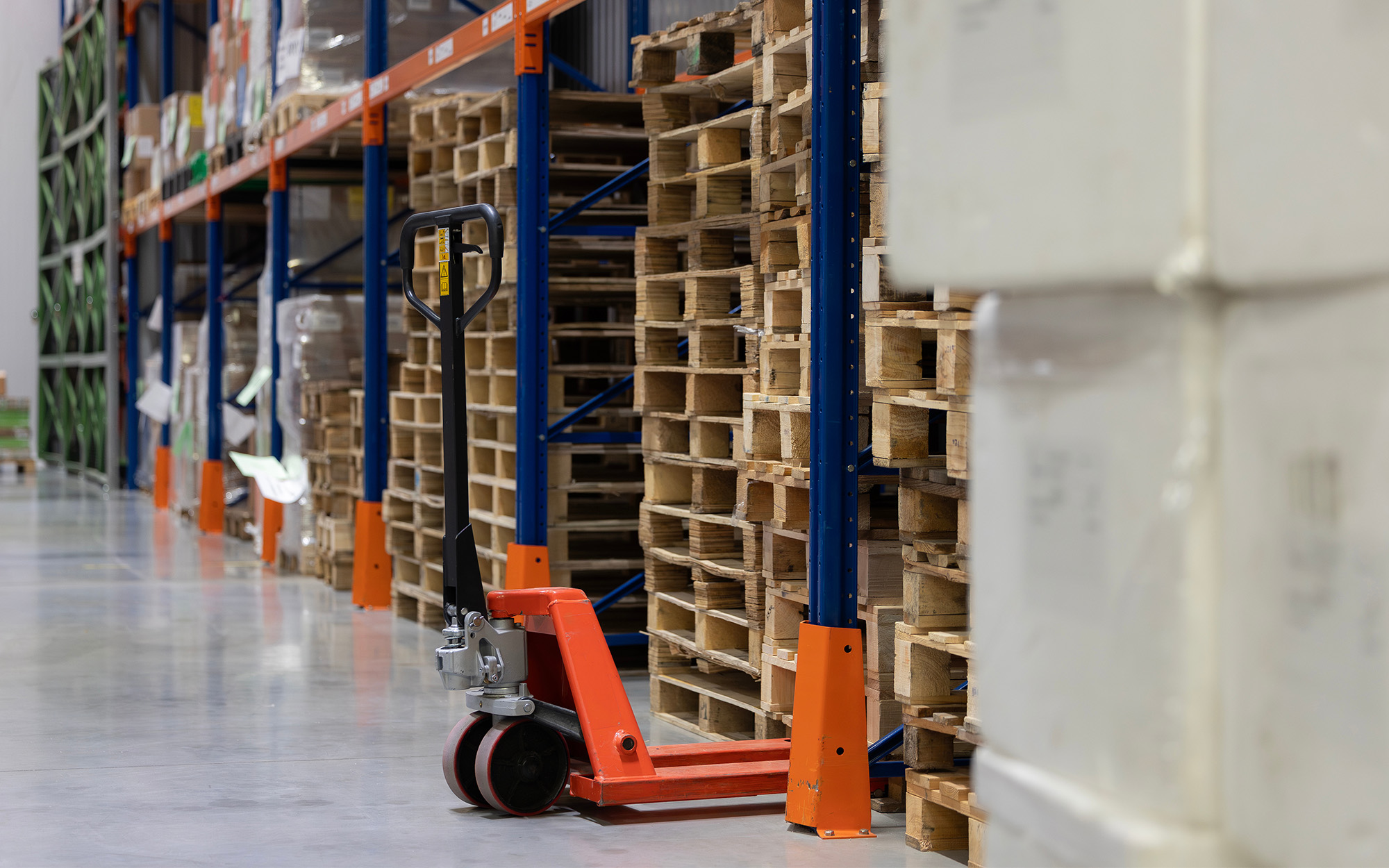 An Orange Manual Pallet Jack Is Parked On A Concrete Floor Next To Stacks Of Empty Wooden Pallets